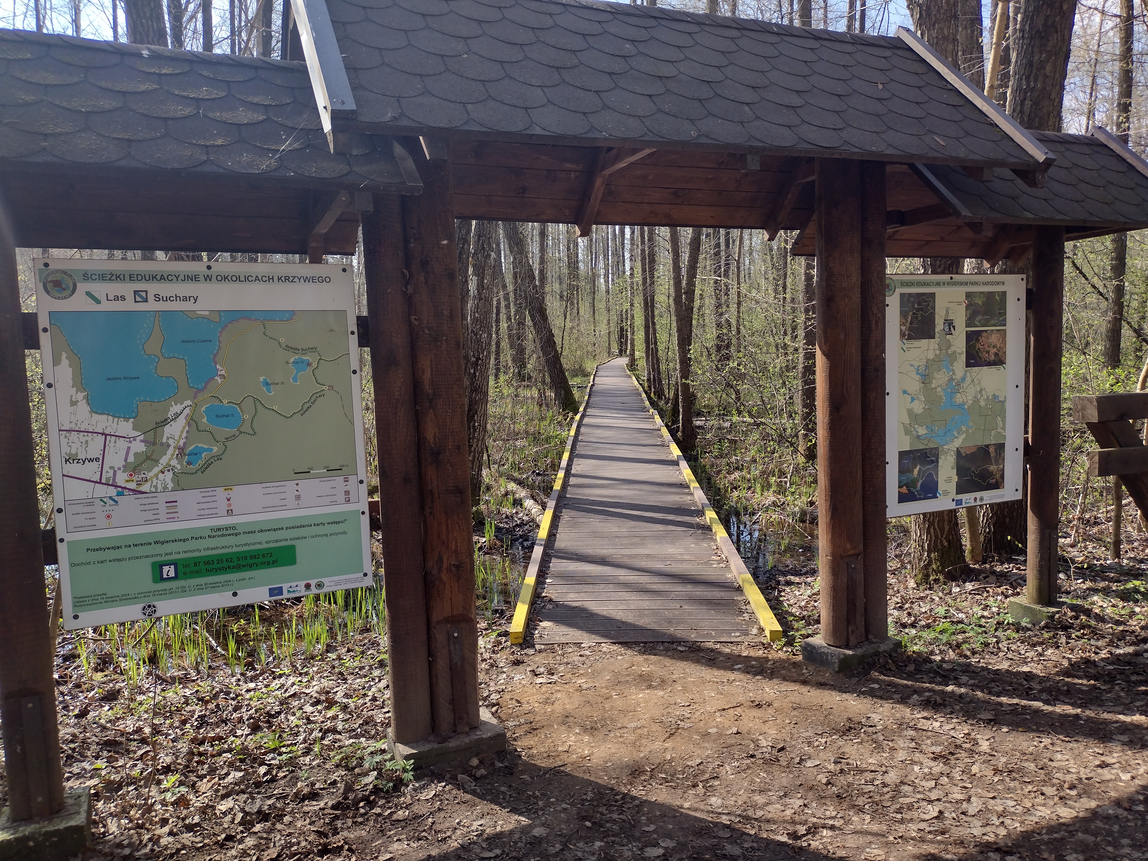Wejście na ścieżkę turystyczną wiodącą przez las. Drewniana brama wejściowa z tablicami informacyjnymi po dwóch stronach. Na tablicach widoczne są mapy. Mapa z lewej strony prezentuje okoliczne ścieżki, z prawej strony mapa całego parku. 