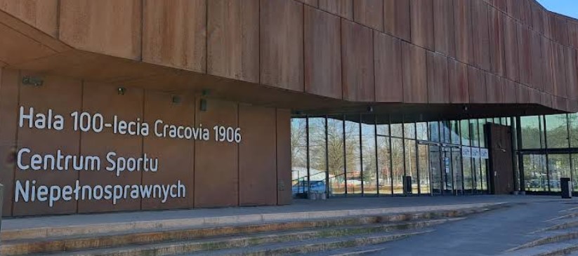 Wejście do nowoczesnego budynku sportowego z przeszkloną elewacją i napisem „Hala 100 lecia Cracovia 1906 – Centrum Sportu Niepełnosprawnych” na ścianie. Przed wejściem schody i podjazd dla wózków.