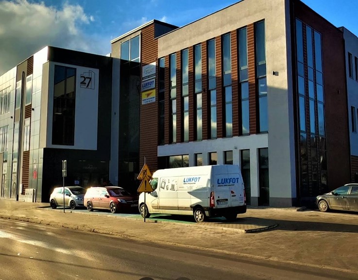 Nowoczesny budynek biurowy z dużymi oknami i jasną elewacją, przed którym stoją zaparkowane samochody wzdłuż ulicy.