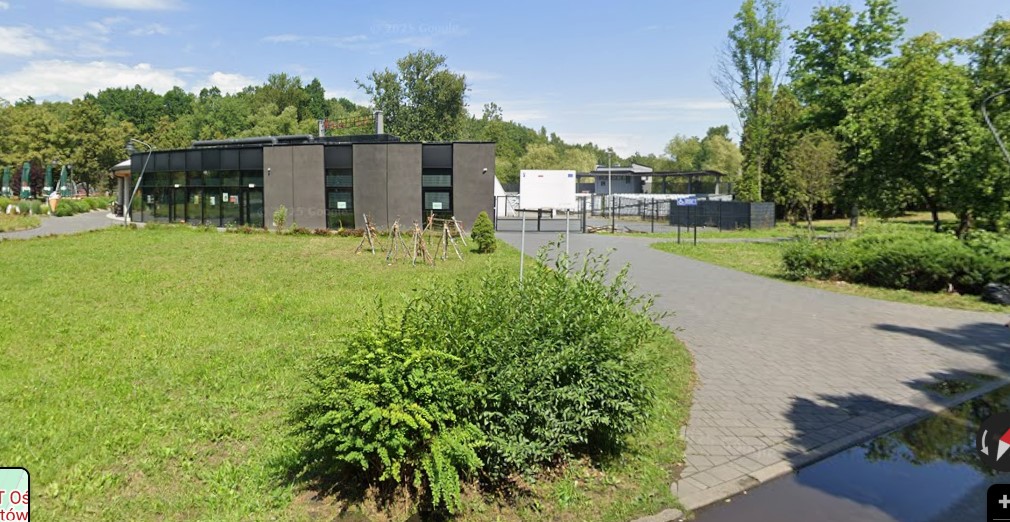 W tle niski, nowoczesny budynek otoczony zielenią, a na pierwszym planie utwardzona ścieżka prowadzącą do wejścia i zielone tereny rekreacyjne wokół.