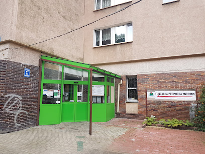 Wejście do budynku z zielonymi oszklonymi drzwiami i zadaszeniem, z tablicą informacyjną „Fundacja Promocja Zdrowia” na ceglanej ścianie obok.