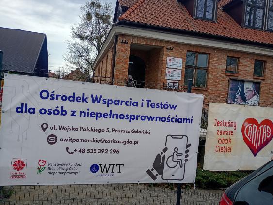 Baner informacyjny na ogrodzeniu przed ceglanym budynkiem z napisem „Ośrodek Wsparcia i Testów dla osób z niepełnosprawnościami”, adresem w Pruszczu Gdańskim, numerem telefonu oraz ikoną dostępności dla osób poruszających się na wózku.