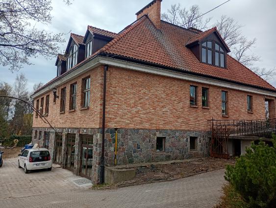 Ceglany, dwukondygnacyjny budynek z czerwonym dachem i kamiennym parterem, widoczny od strony podjazdu z zaparkowanym samochodem oraz drzewami w tle.