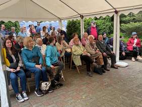 Pokaż zdjęcie: Grupa uczestników spotkania siedzi na krzesłach pod dużym namiotem z odkrytymi ściankami