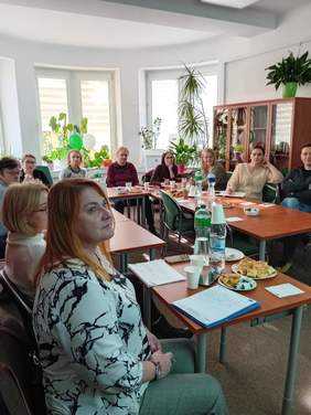 Pokaż zdjęcie: Grupa osób siedzących na krzesłach za stołem w sali z uwagą słuchają prelegenta