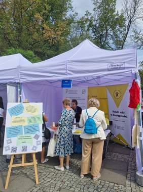 Pokaż zdjęcie: Na świeżym powietrzu stoi biały namiot, w którym jest stolik z materiałami PFRON, w tle stoją 3 roll upy z logotypami PFRON. Przy stoisku widać dwie osoby, które przeglądają materiały. Rozmawiają między sobą i z kobietą obsługującą stoisko. Na pierwszym planie sztaluga z planszą, na której są hasła promujące dostępność.