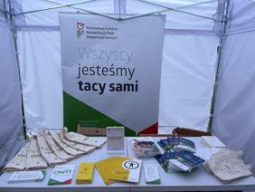Pokaż zdjęcie: Pod namiotem na stole znajdują się materiały promocyjne, w tle roll-up PFRON