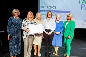 Pokaż zdjęcie: Sześć kobiet elegancko ubranych stoi na scenie. Dwie z nich trzymają w rękach dyplom z podziękowaniami. 