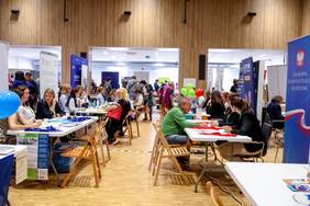 Pokaż zdjęcie: W dużej chali znajdują się boksy informacyjne wystawców. Przed każdym z nich reprezentanci instytucji przy stolikach udzielają informacji.