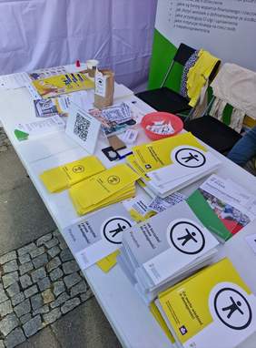 Pokaż zdjęcie: Na białym stoliku leżą materiały PFRON o dostępności: broszury, poradniki i ulotki. Są w kolorach białym i żółtym, stoi też pudełko z cukierkami, tabliczka z kodem QR.