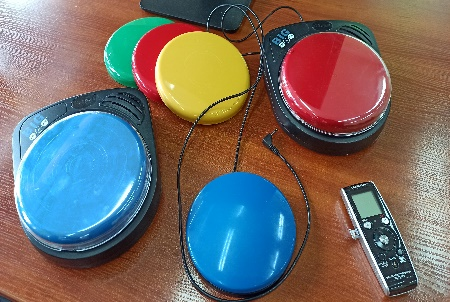 Pokaż zdjęcie: Na stole znajduje się 6 kolorowych, okrągłych specjalistycznych przycisków oraz prostokątny dyktafon