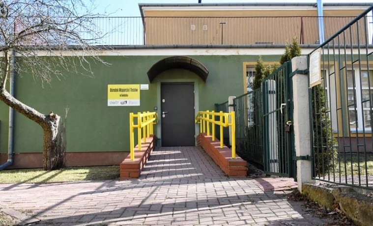 Pokaż zdjęcie: Wejście do zielonego budynku wraz z podjazdem i żółtymi poręczami, po prawej stronie zdjęcia ogrodzenie