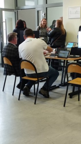 Pokaż zdjęcie: Przy stoliku siedzi dwóch mężczyzn. Osoba w koszulce prezentuje technologie OWiT. W tle grupa kobiet dyskutuje o wystawionych urządzeniach.